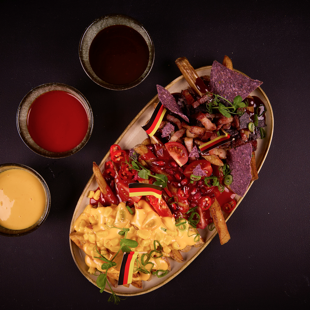 Aufgetürmte selbst frittierte Pommes auf einem Teller, belegt mit drei Saucenzonen aus BBQ Bier Dip mit Bacon-Crunch, Fire Cracker Sauce mit Chili und Granatapfel sowie Melted Cheese Dip mit Mais, Nacho Chips und Frühlingslauch